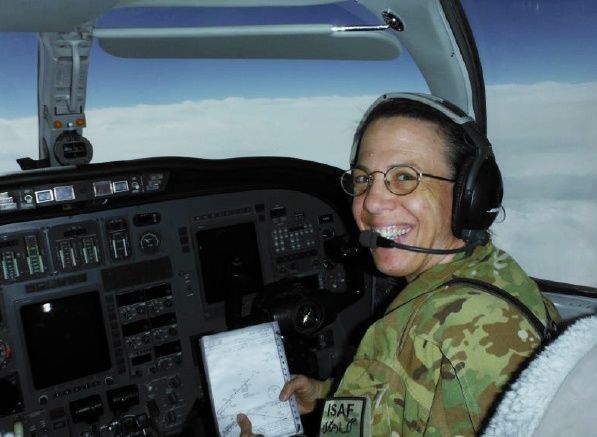 Fondy, dressed in camo, smiles from the cockpit of a military aircraft and holds up a notebook. Out the window, blue sky and a layer of clouds seen from above.