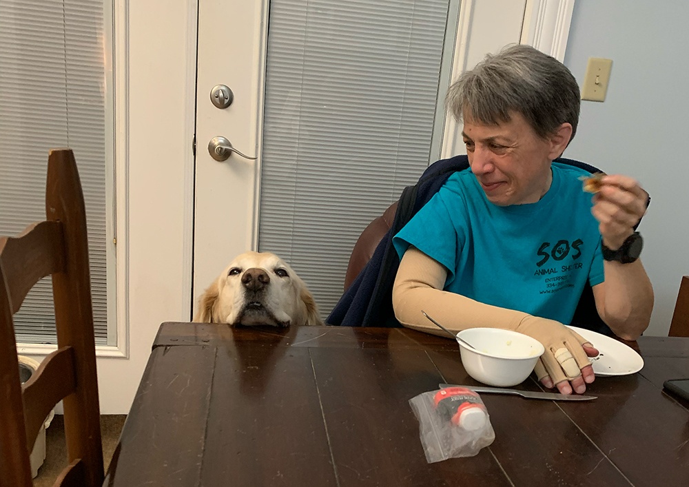 Fondy sits eating, smiling down at the head of a golden retriever popping up over the edge of the table. Fondy's lymphedema garment is visible on her right hand and arm.