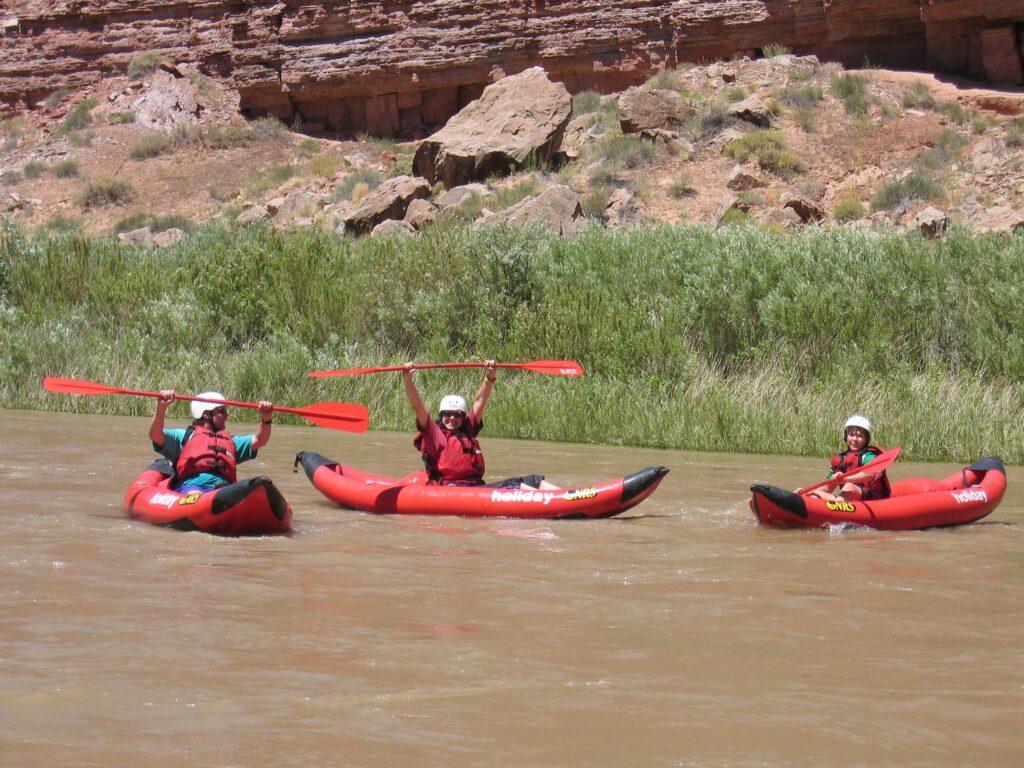 Beckerle and her family in three inflatable kayaks on calm river water. Beckerle raises her paddle above her head, smiling. The red rocks of Utah are seen in the background.