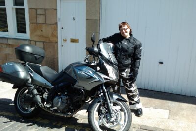 Andi smiles next to a motorcycle, in full leather gear, holding a helmet.