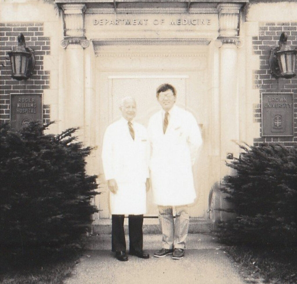 Visibly aged photo of Calabresi and Chu posing in white coats outside a stone building with engraved "School of Medicine" archway.