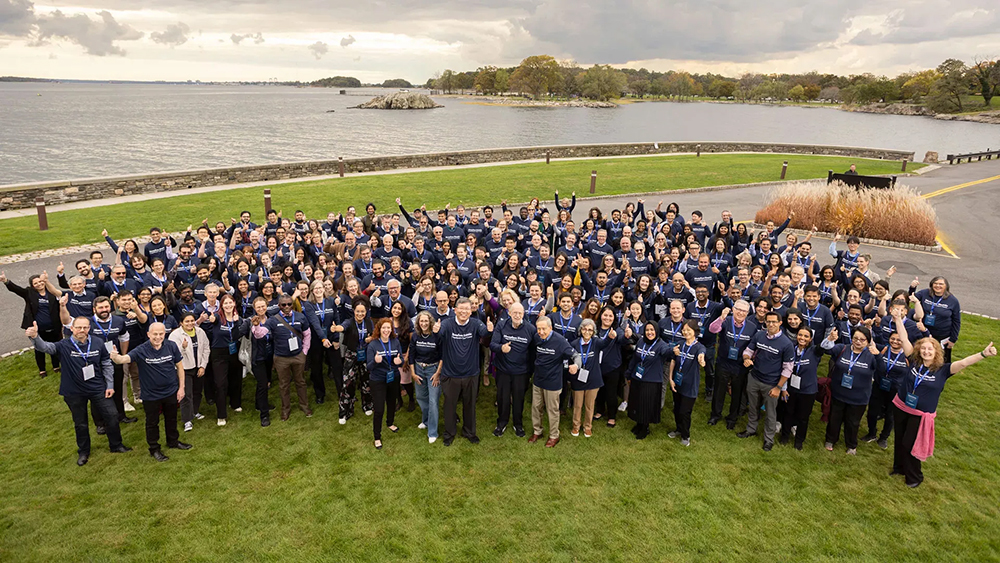 Chu holds two thumbs up, beaming, at the head of a huge group of people in matching shirts posing on a waterfront lawn. 