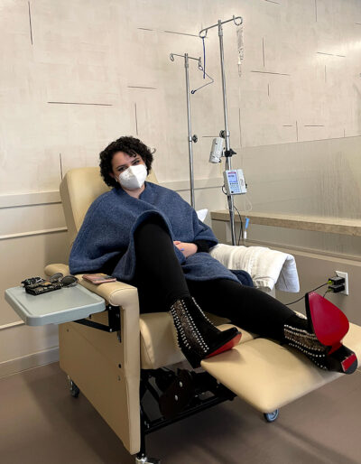Sandino sits in an infusion chair in red-bottomed, metal-spiked, high-heeled boots.
