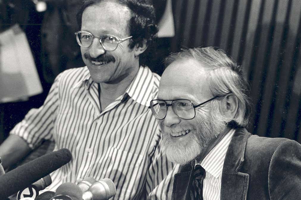 Black and white photo of Varmus and Bishop smiling while sitting in front of microphones at a press event.