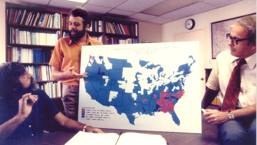 Aged photograph showing Hoover and Mason in discussion as Fraumeni looks on. Hoover props up a poster board of cancer mortality rates across the U.S. 