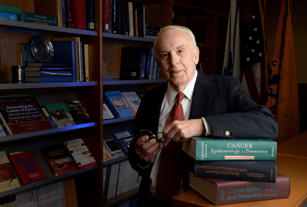 Fraumeni poses in a library, leaning on a pile of "Cancer Epidemiology and Prevention" textbooks written by him and David Schottenfeld.