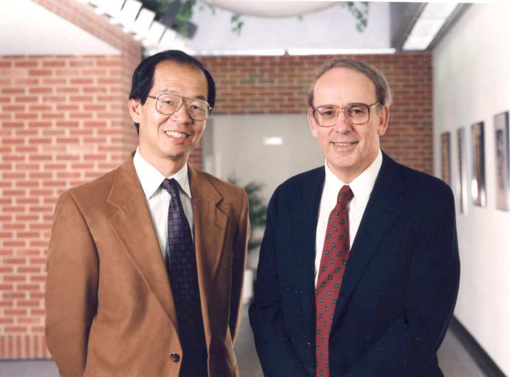 Li and Fraumeni smile together in suits, standing in a hallway.