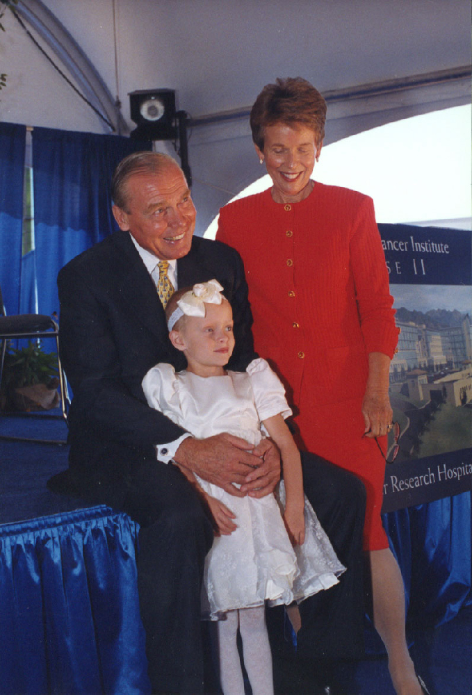 Karen and Jon pose with a pediatric cancer patient who is wearing a white dress and headband. 