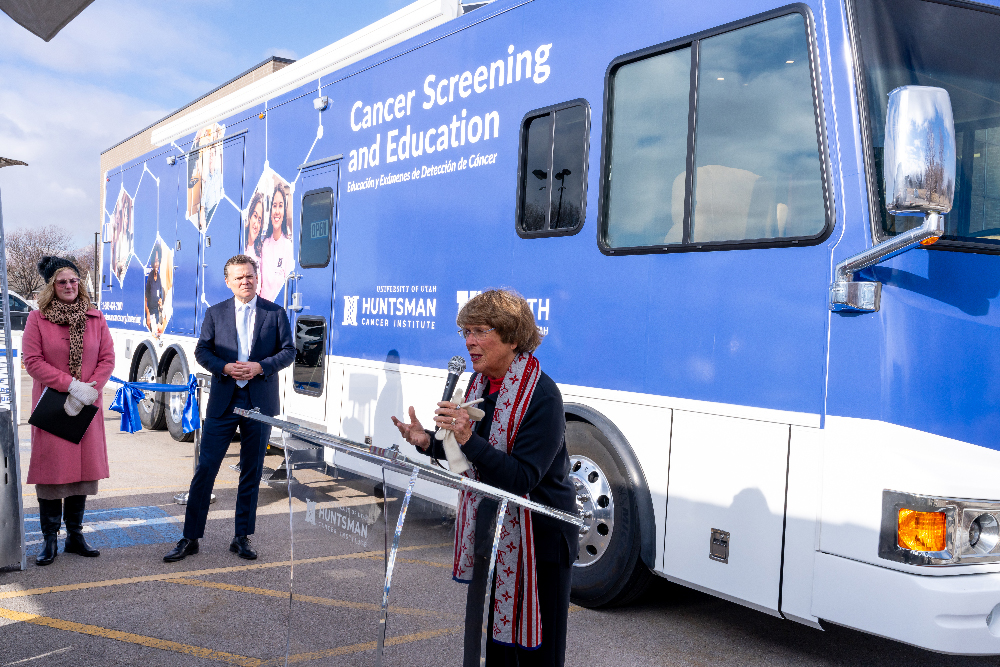 Karen speaks at a podium in front of a large Huntsman mobile cancer screening vehicle. 