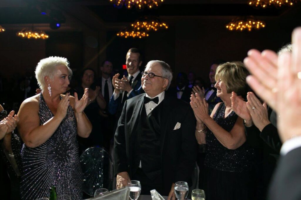 Morrow, in formal wear and a bowtie, stands in a spotlight surrounded by people giving him a standing ovation and applauding him.