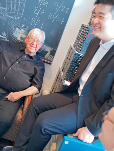 Dr. Szalay sits in a chair in front of a chalkboard, Lu laughs in the foreground.