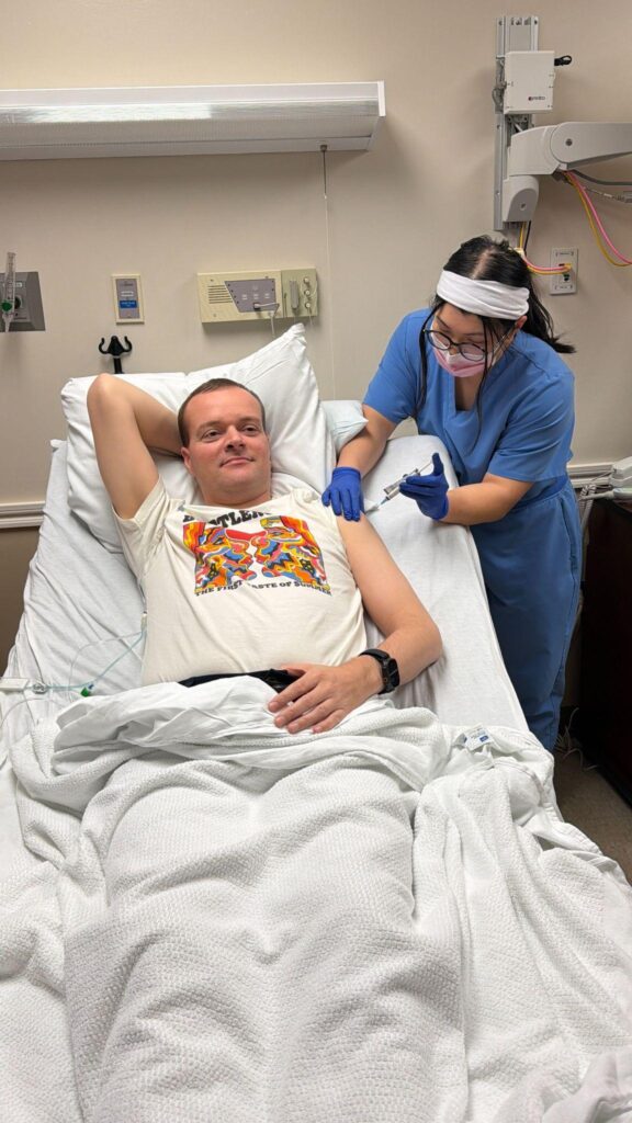 Sijbrandij, lying in a hospital bed, receives injection from a clinician.