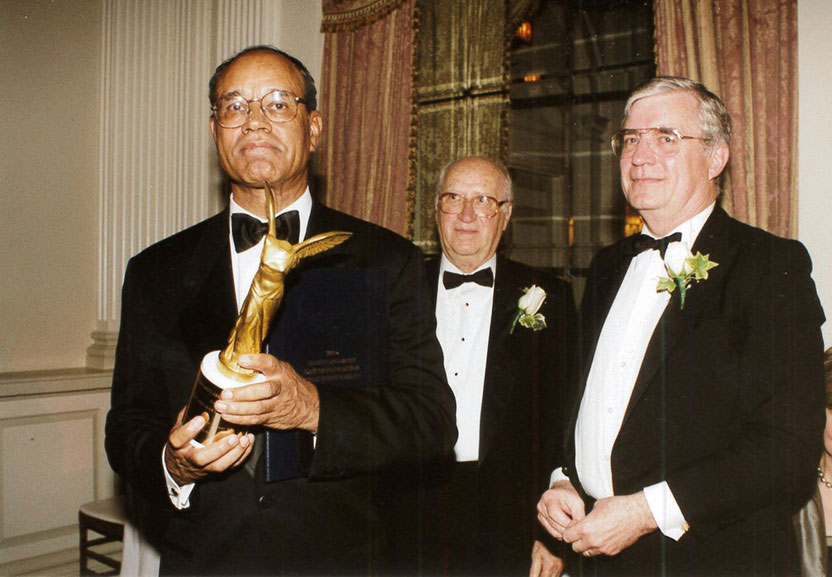 Freeman in a tuxedo holds a golden Lasker award, with Koshland and Fordyce behind him, also in tuxedos.