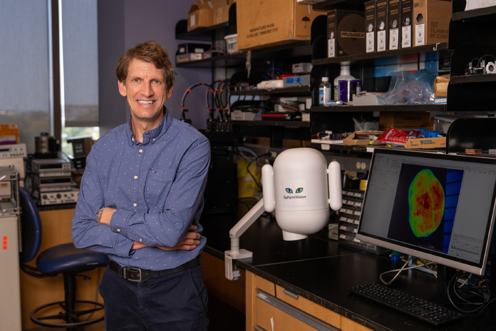 Haemmerich in the lab, next to a small white oblong device with a decal of cat eyes, on a swing arm.
